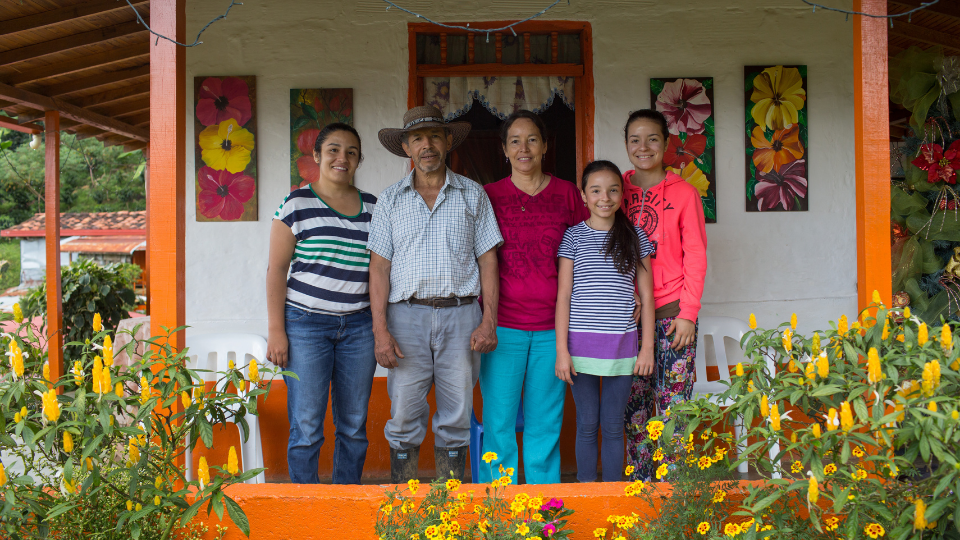 A third generation coffee farmer in Colombia poses for a photo in front of his brightly colored home alongside his wife and three daughters.