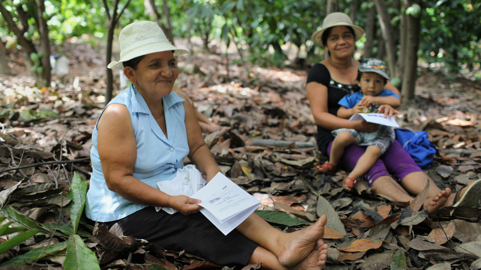 Two women sit on the ground under cacao trees; one woman holds her baby in her lap.